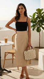 Woman in black top and beige skirt holding a woven handbag on a balcony.