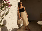 Woman holding a woven bag against a white wall with greenery