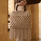 Beige woven handbag with fringe held by a person in a blurred indoor setting