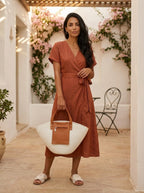 Model standing in courtyard with wooden pergola holding the Soleil Tote at her side, terracotta wrap dress with cognac leather sandals