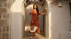 Woman in terracotta linen wrap dress carrying the Soleil Tote through a whitewashed stone archway with bougainvillea, golden hour light